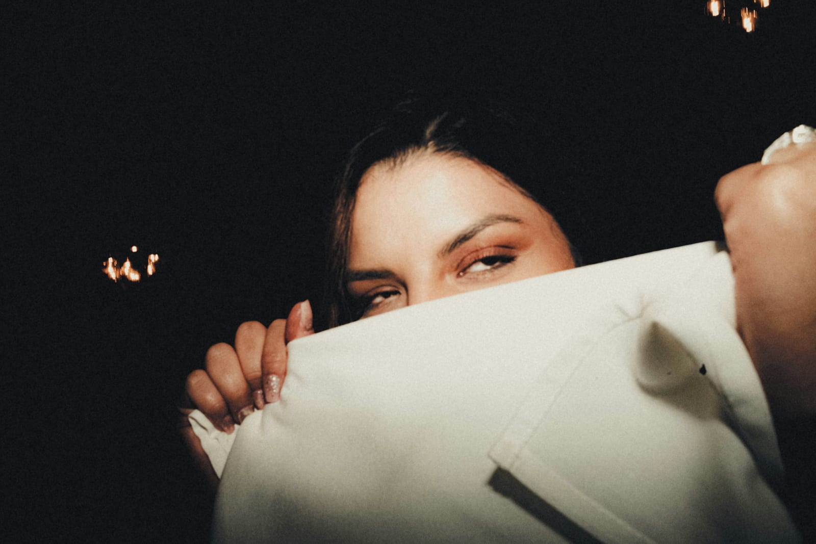 Close-up of a woman peeking over a white napkin in a dark room, eyes half-closed, with warm lights and a sleepy mood