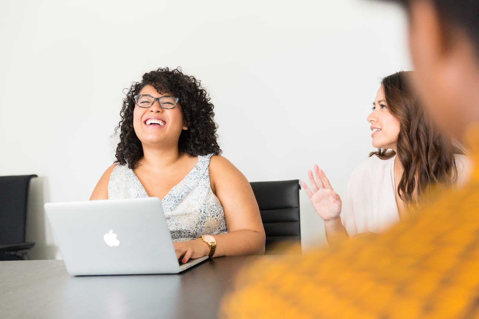 Two women in an office setting, one laughing with a laptop, the other gesturing. Bright, cheerful mood. Minimal, white background.