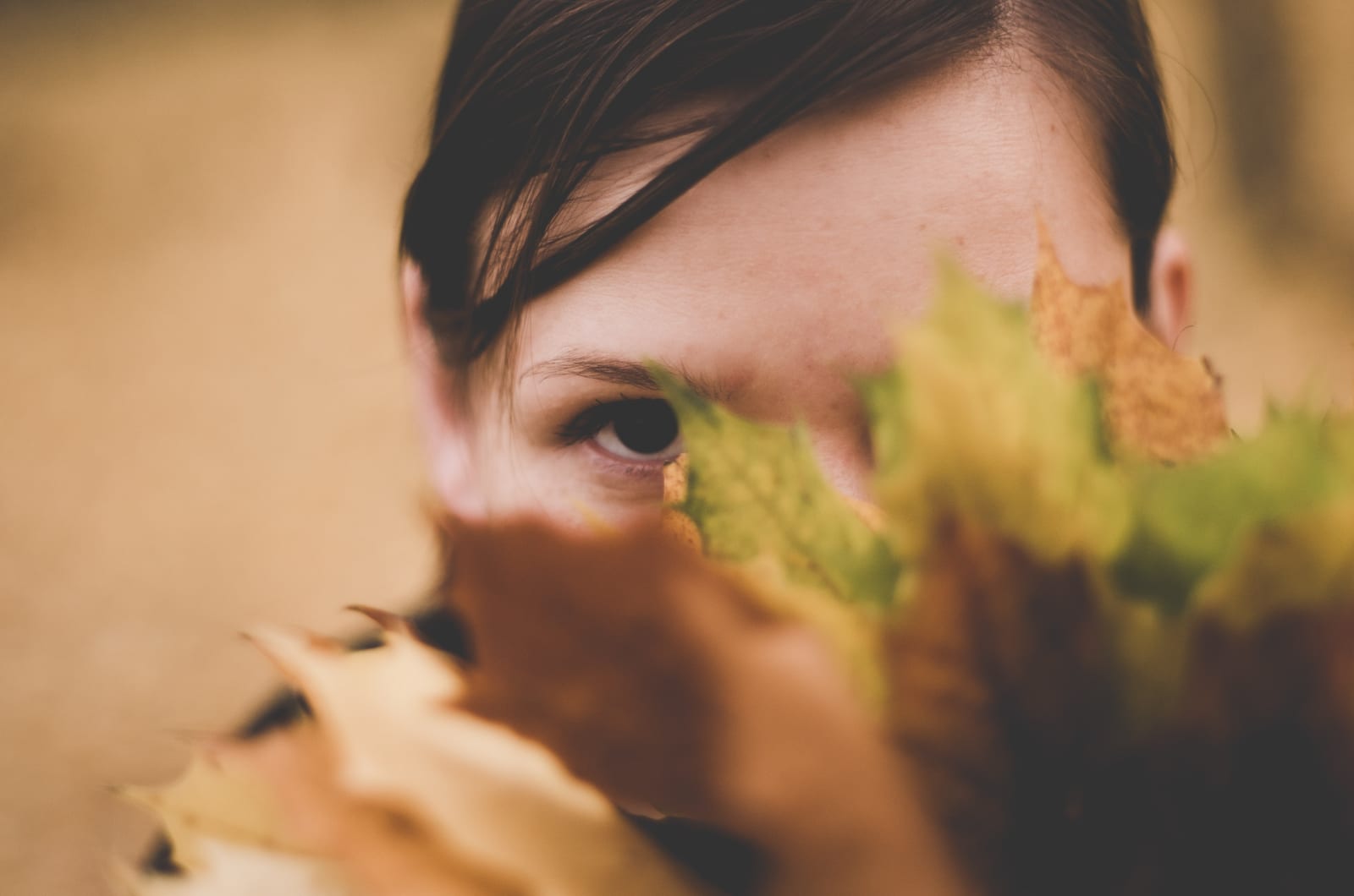 Face partially hidden by autumn leaves, with one eye visible. Blurred background adds focus to the warm tones and mysterious mood.