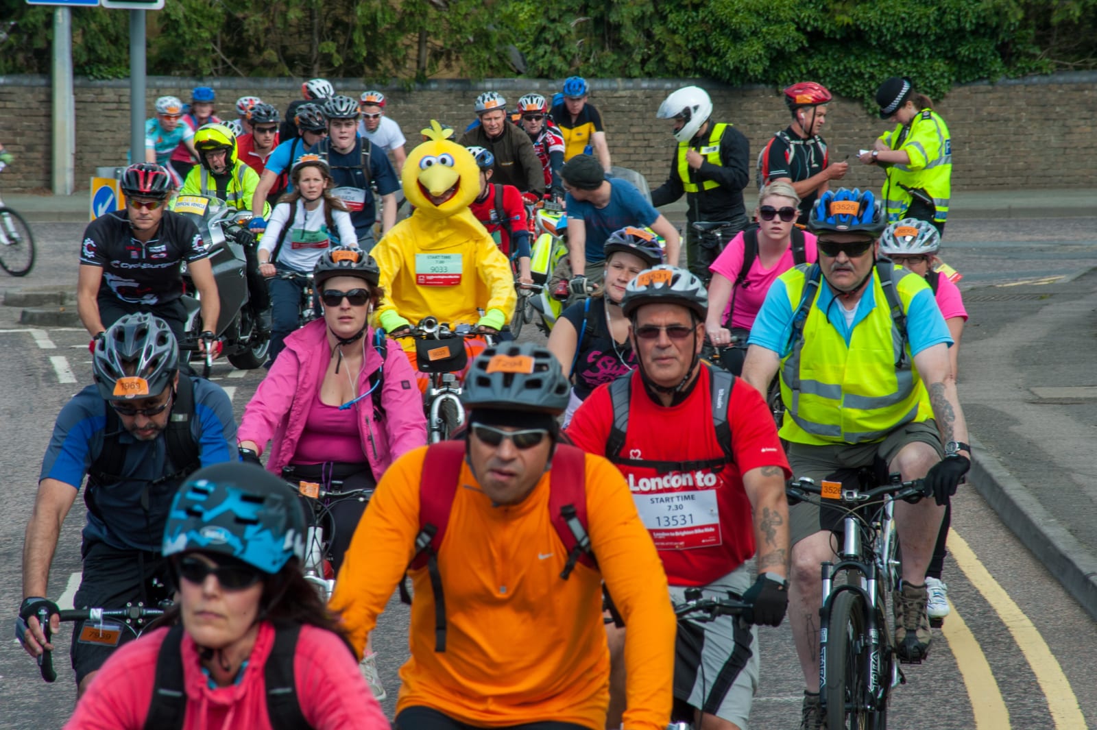 Cyclists in colorful gear, including one in a yellow bird costume, ride on a city street. Police and spectators watch from the background.