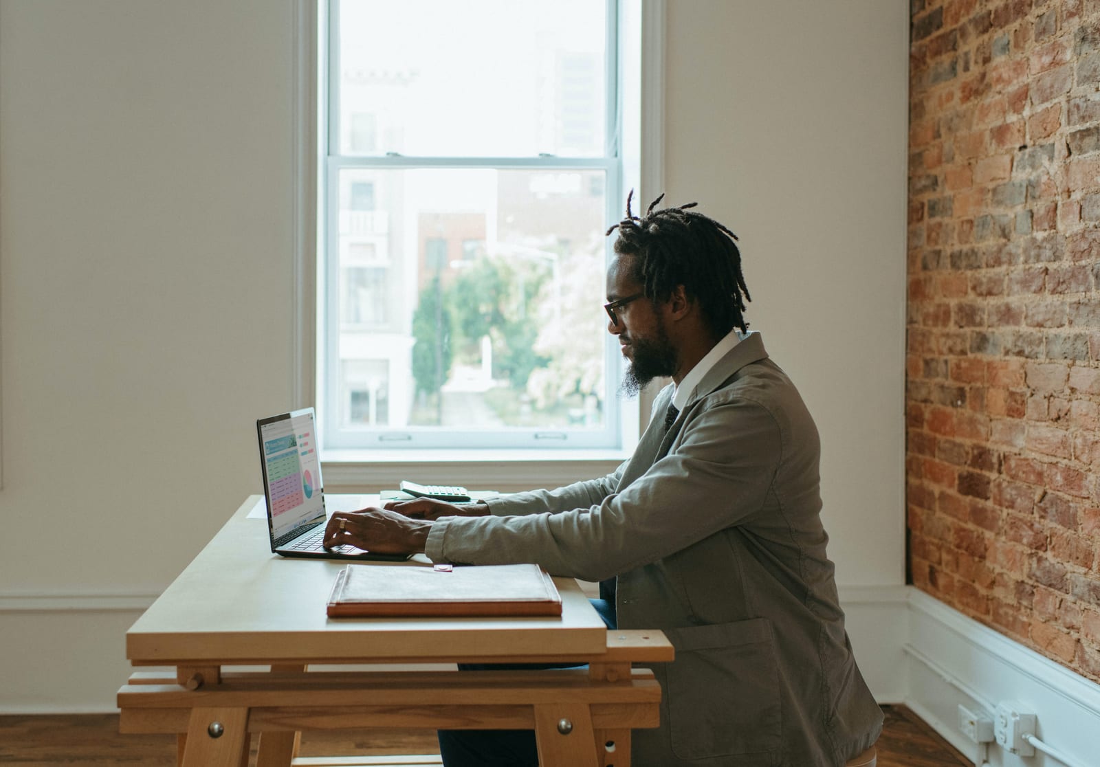 Man in a suit works on a laptop at a wooden table in a bright room with an exposed brick wall and large window showing a city view.