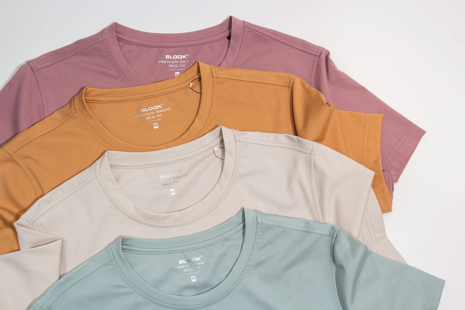 Four neatly stacked T-shirts in pink, mustard, beige, and teal. Each shirt has "BLOOK PREMIUM BRAND" printed on the chest.