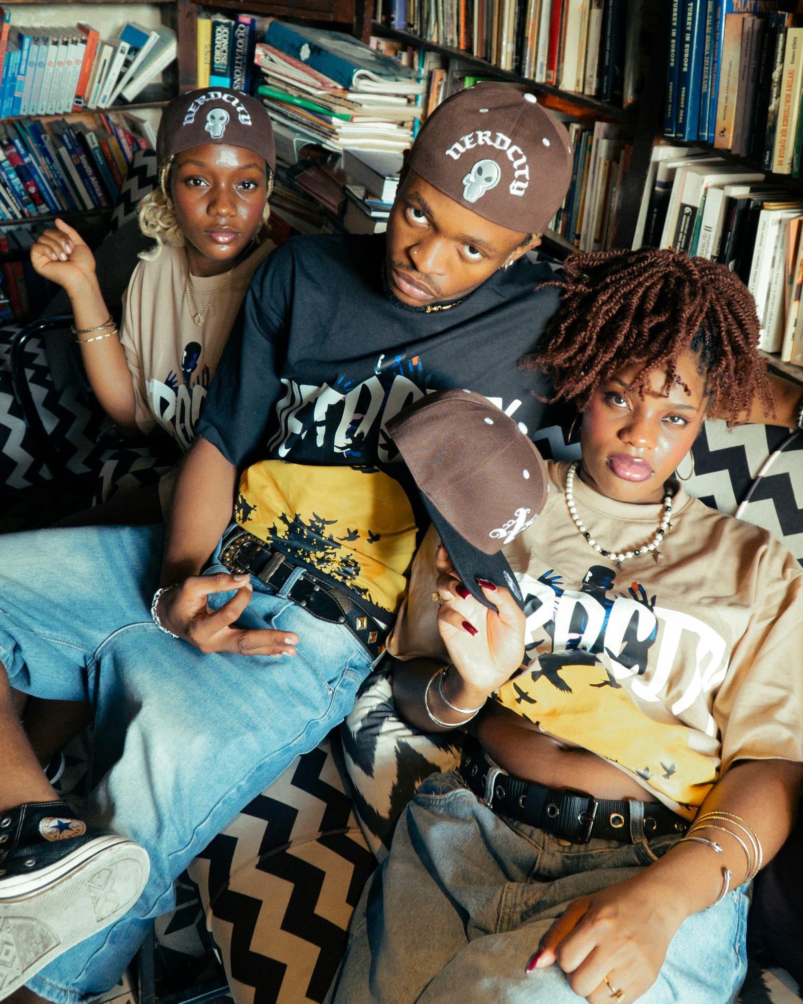 Three people in casual wear sit on a patterned couch in a library, wearing matching caps. They convey a relaxed, stylish vibe.